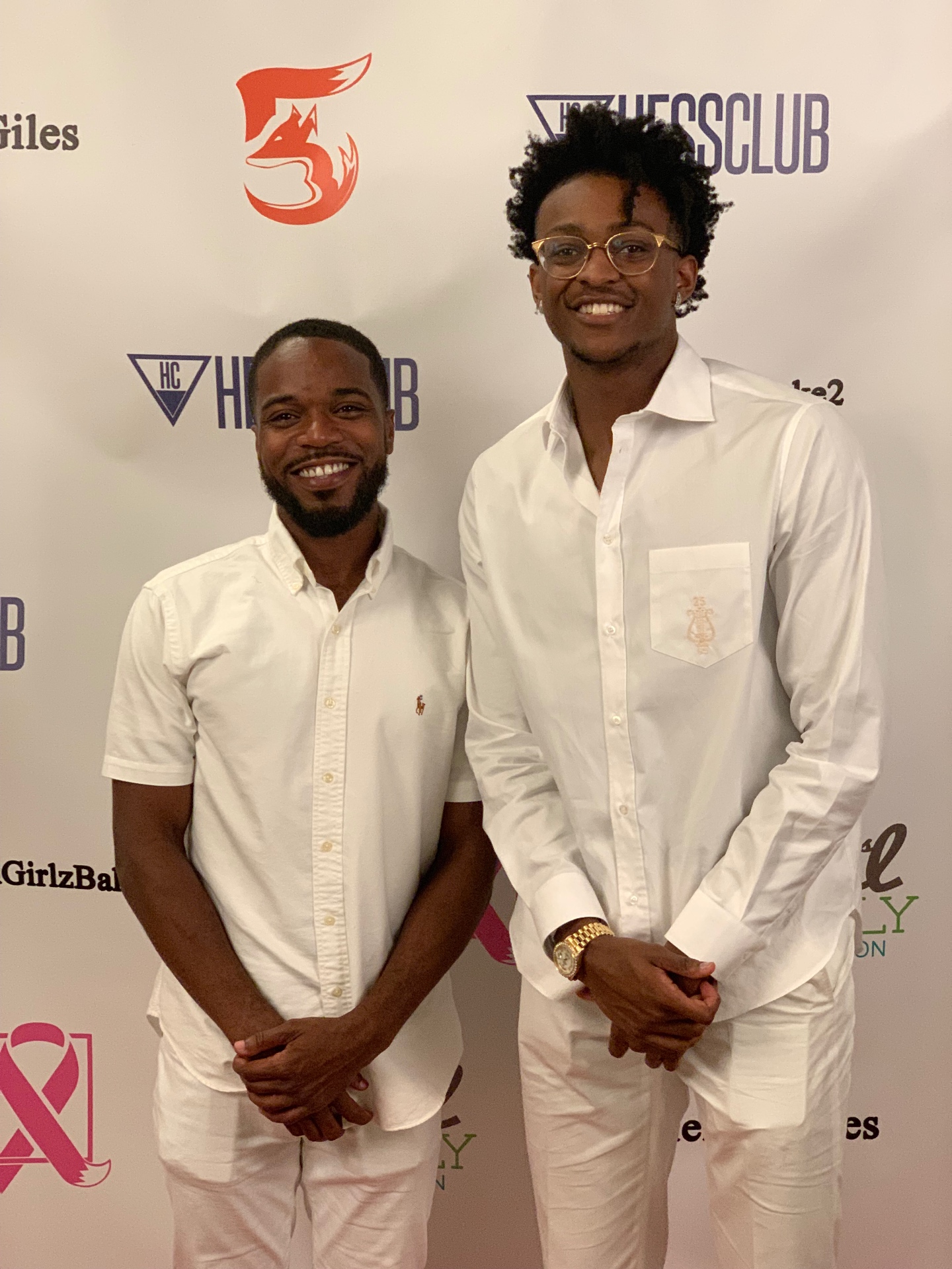 Two men in white outfits standing together in front of a charity event backdrop.