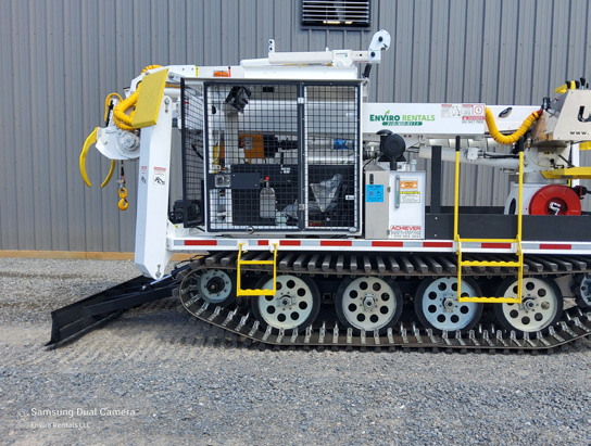 Tracked industrial equipment with white body panels and yellow fittings parked beside a gray building.