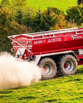 Red restoration equipment trailer dispersing material beside green grass and trees.